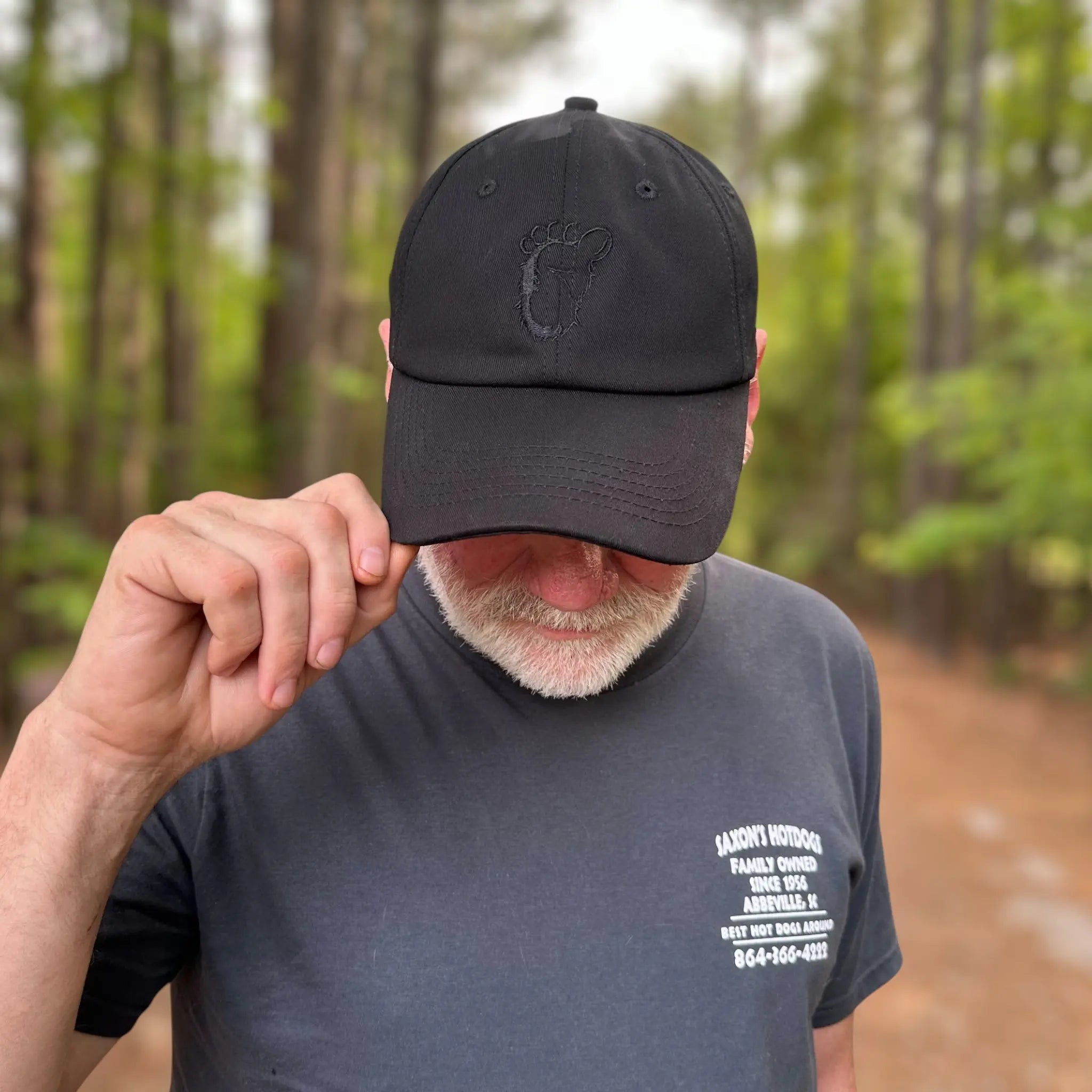 Man wearing the black Stealth Pine Dad Hat on a forest trail, lightly gripping the brim. The hat’s sleek, understated design is ideal for nature lovers and off-grid souls.