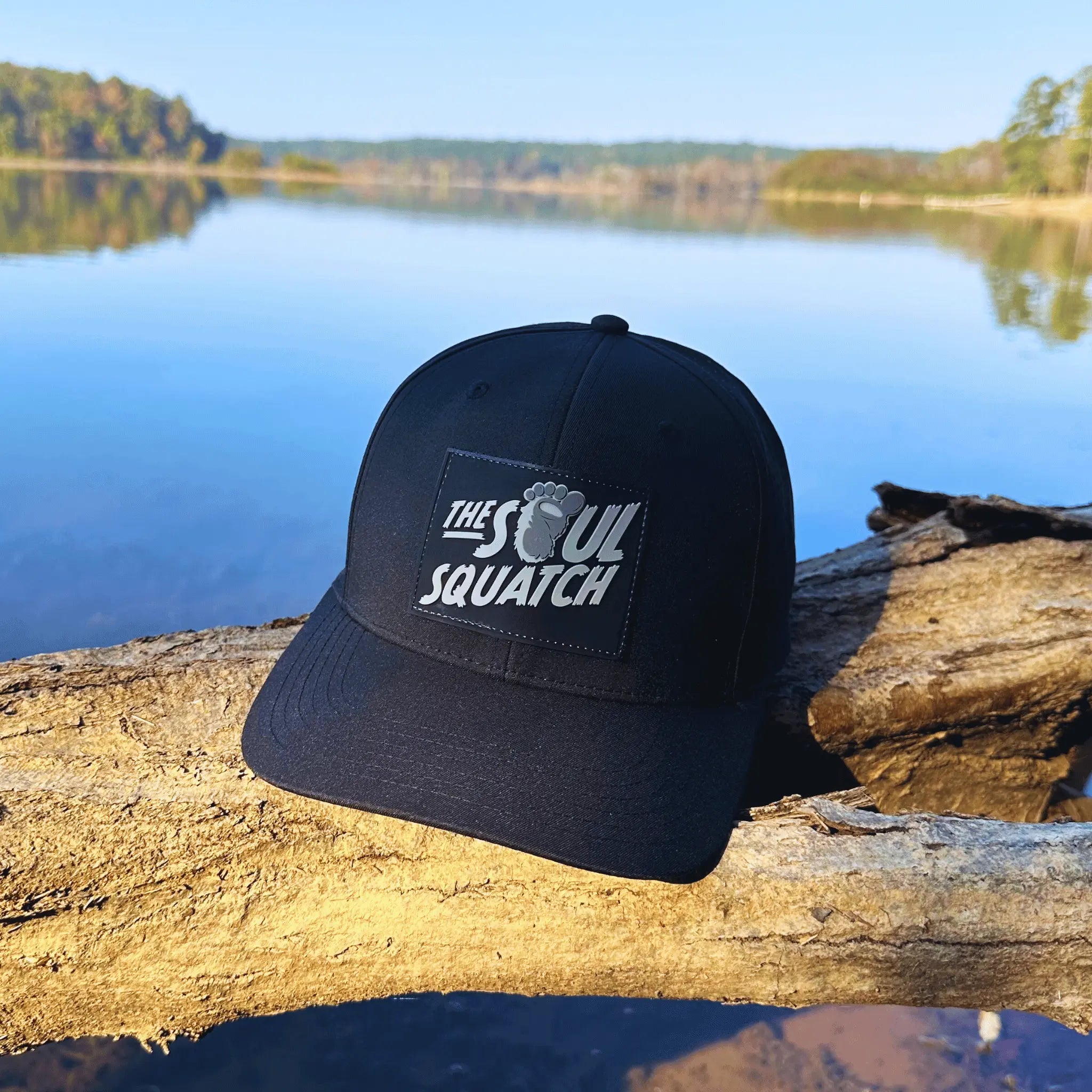 Black Pathfinder Mid-Profile Hat by The Soul Squatch resting on a lakeside log – perfect trail companion for outdoor explorers and Squatch seekers.