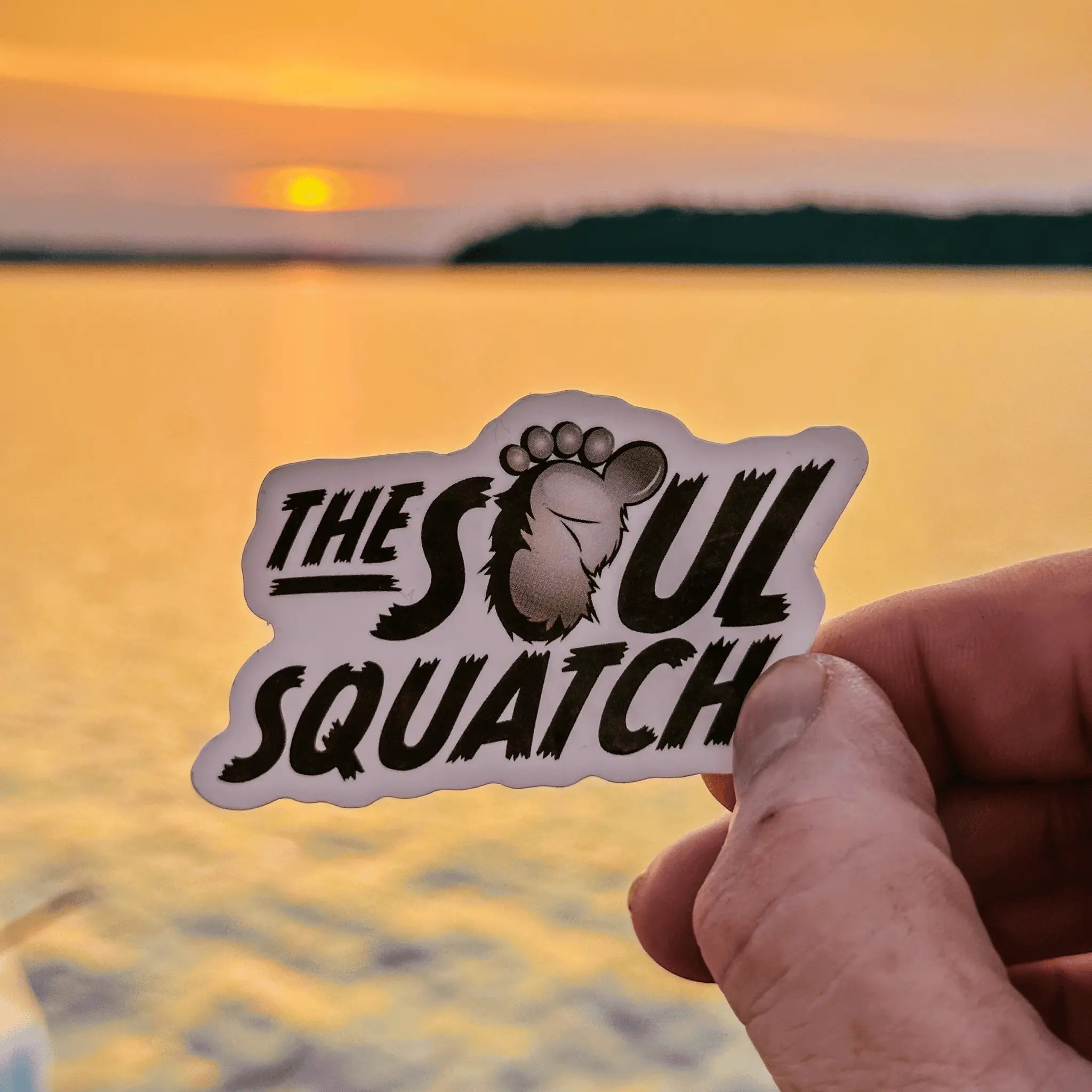 Hand holding The Soul Squatch logo sticker in front of a lake at sunset — weatherproof vinyl sticker with Bigfoot footprint logo, ideal for water bottles and gear.