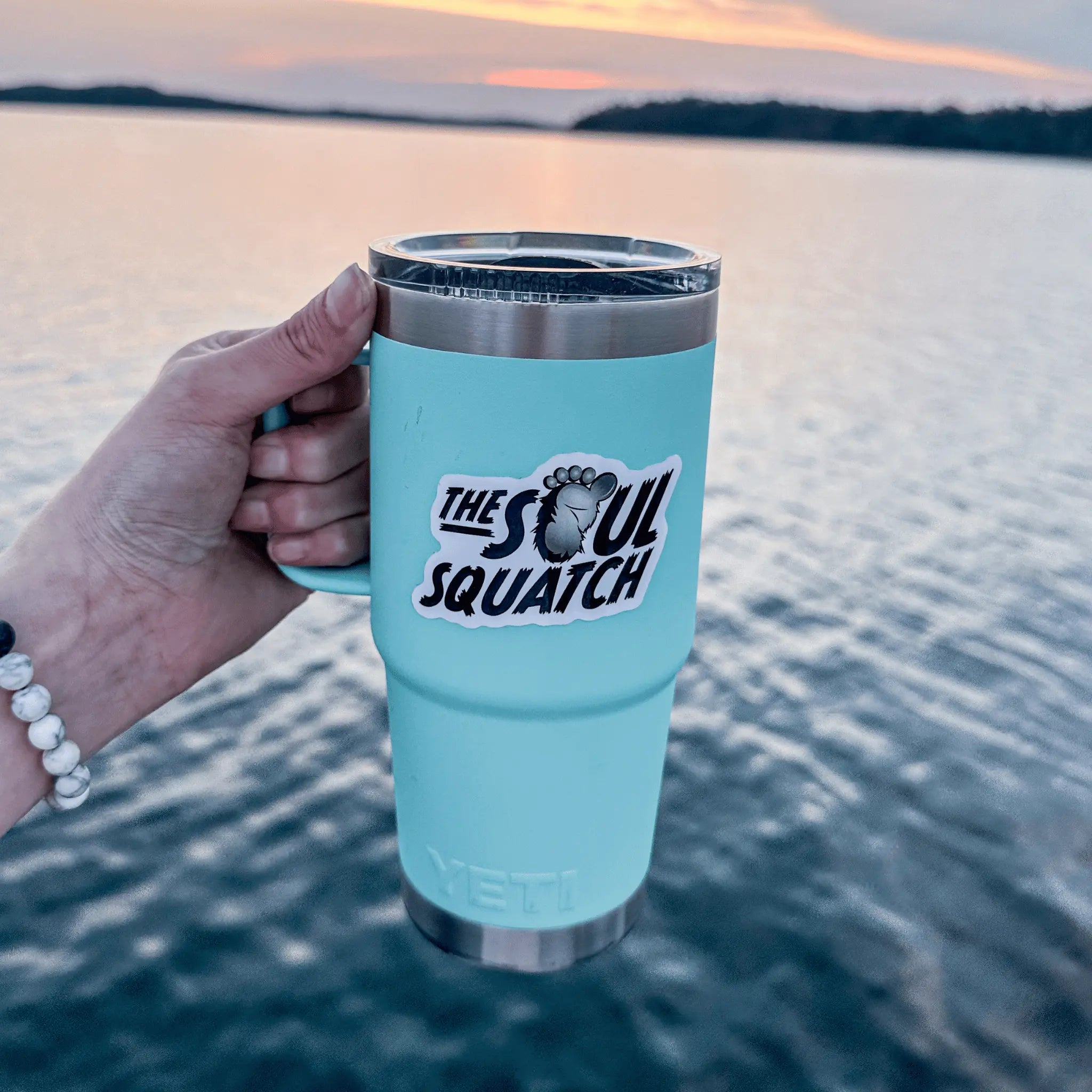 Hand holding a light blue YETI tumbler with The Soul Squatch logo sticker by a lake at sunset — waterproof Bigfoot vinyl sticker perfect for outdoor gear.