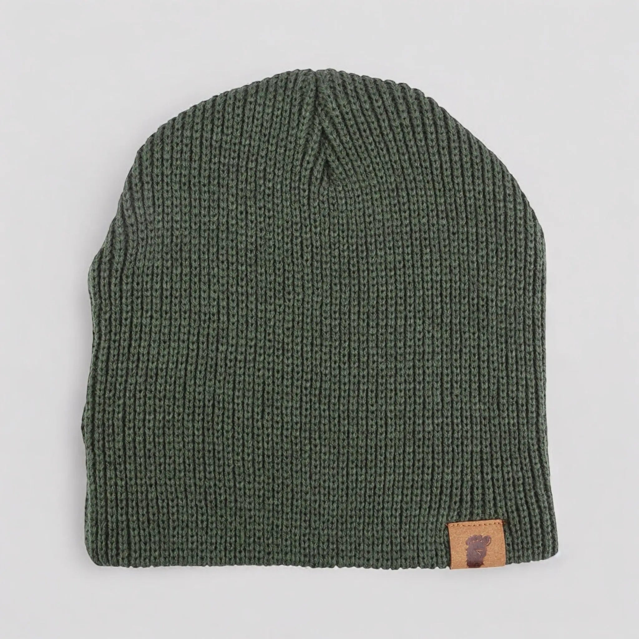 Flat lay of the Wilderness Wanderer Beanie in Skunk Ape Green — ribbed forest-green knit beanie with leather Bigfoot footprint patch.