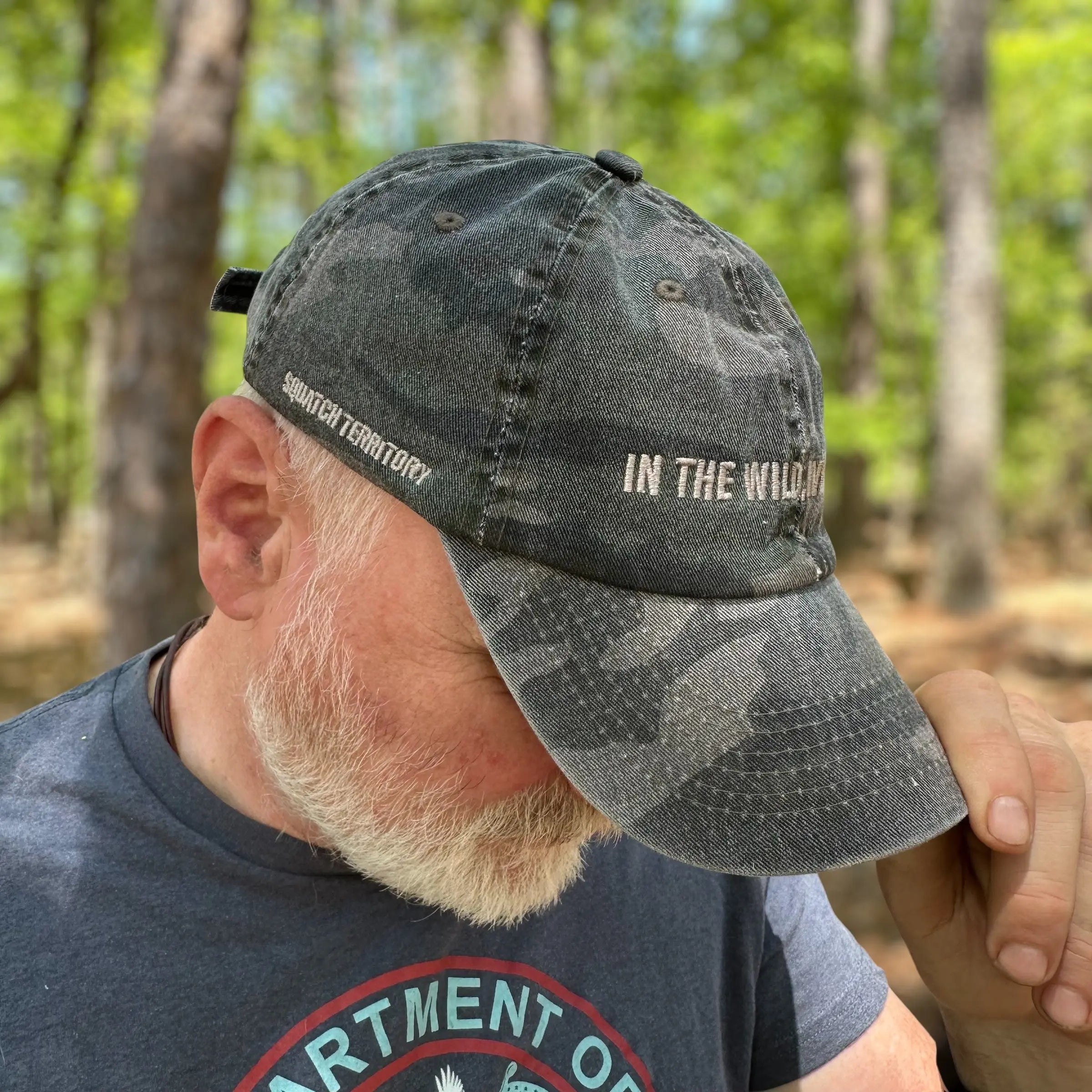 Overhead view of bearded man adjusting The Soul Squatch camo dad hat with outdoor-themed shirt; represents rugged, trail-tested Soul Squatch style.