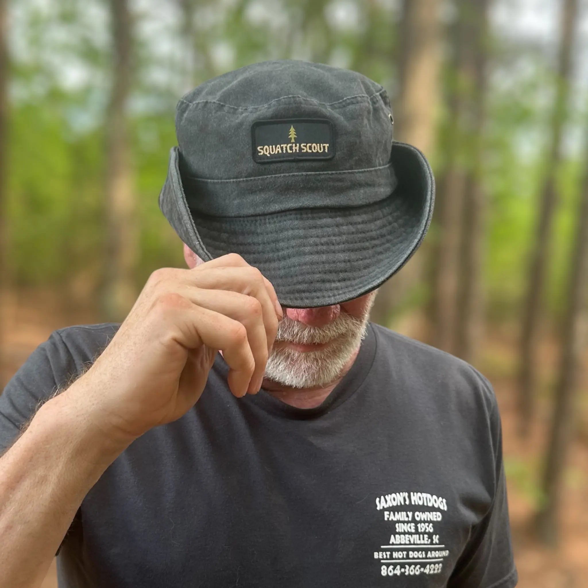 Man tipping the Squatch Scout Boonie Hat on a wooded trail, showcasing the hat's wide brim, vintage finish, and secure chin strap—ideal for adventure-ready souls who roam deep into Sasquatch country.