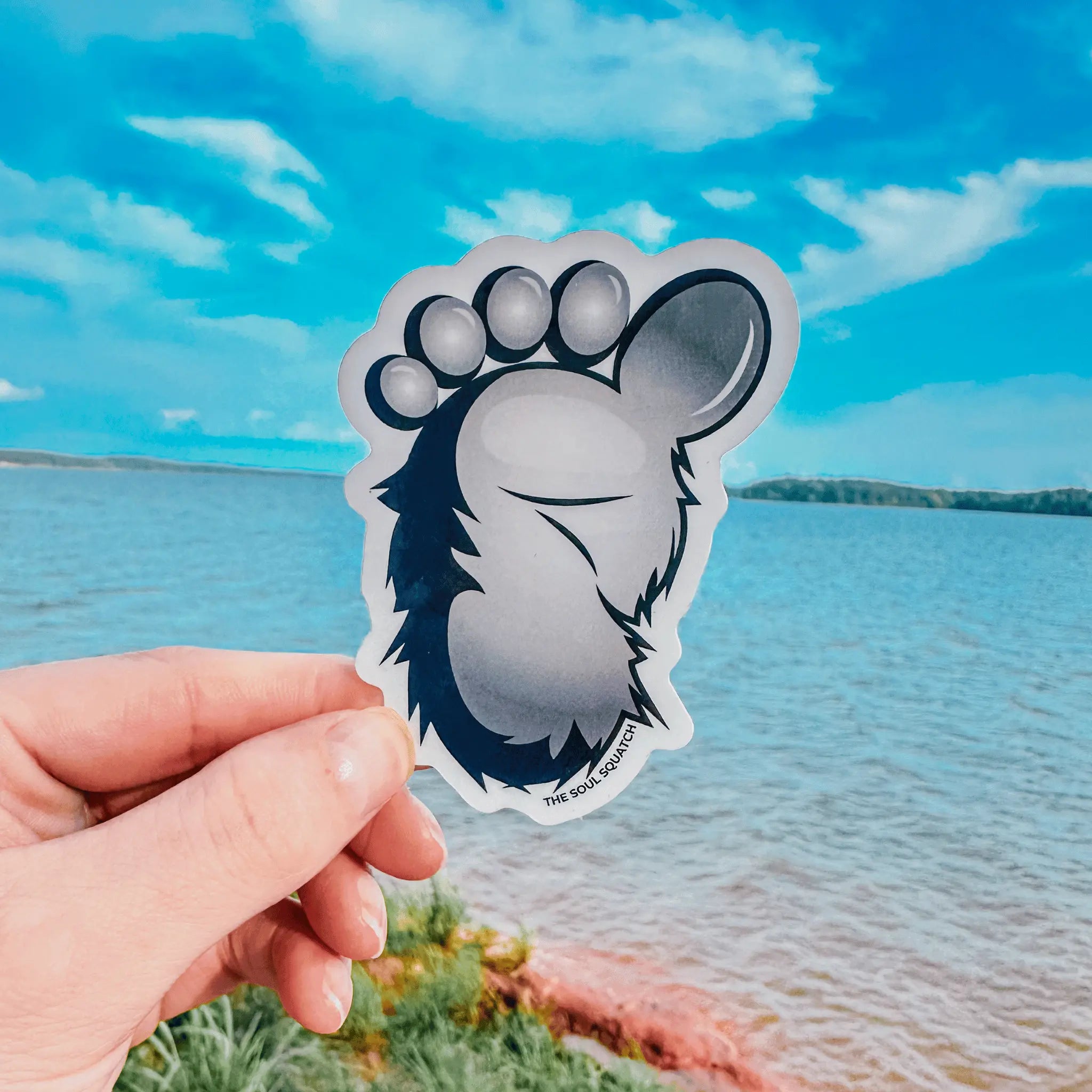 Hand holding the Squatch Footprint Sticker by a lake under a blue sky — durable Bigfoot foot decal designed for water bottles, coolers, and trail-tested gear.