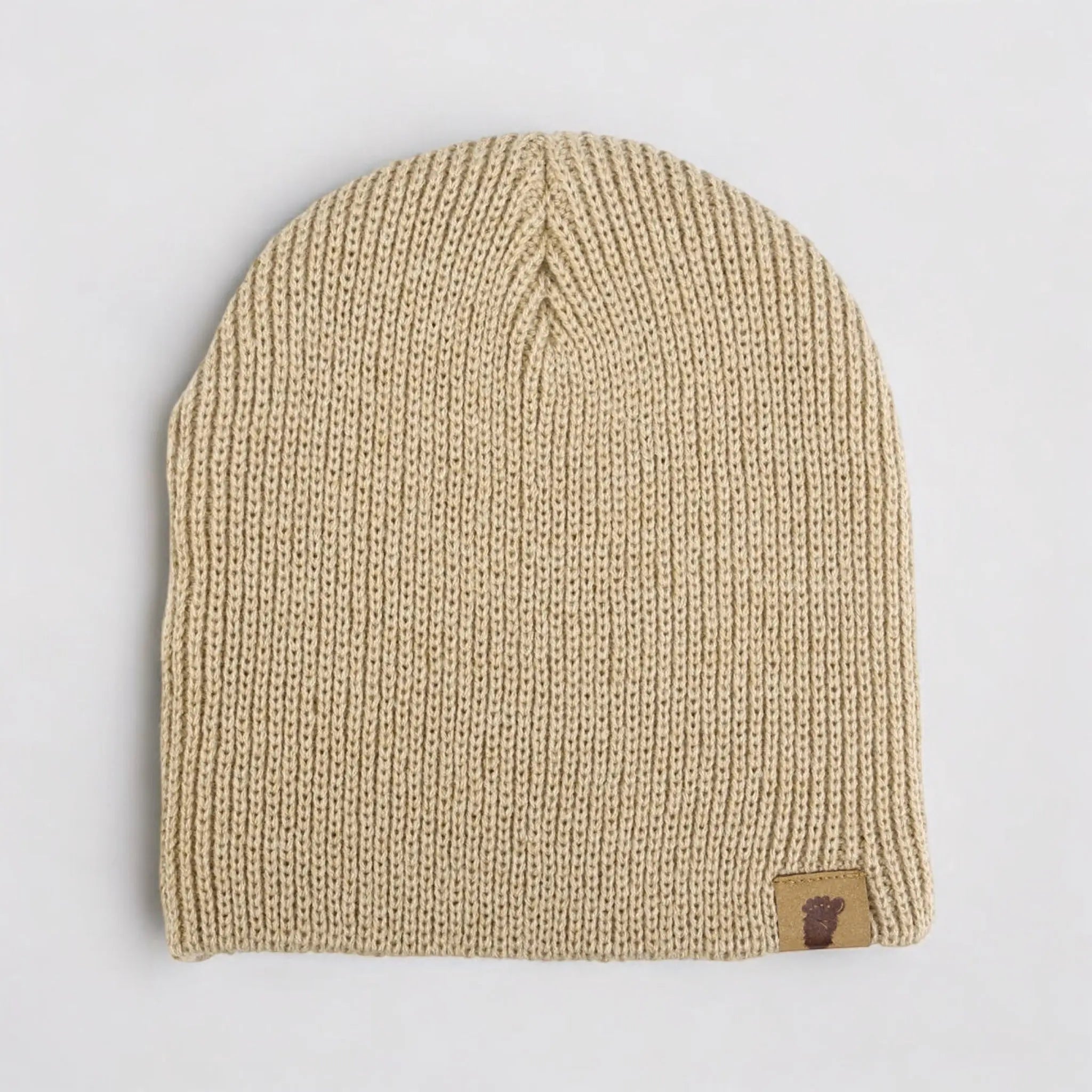 Flat lay of the Wilderness Wanderer Beanie in Skookum Dirt Tan — neutral ribbed tan beanie with leather  Sasquatch footprint patch.