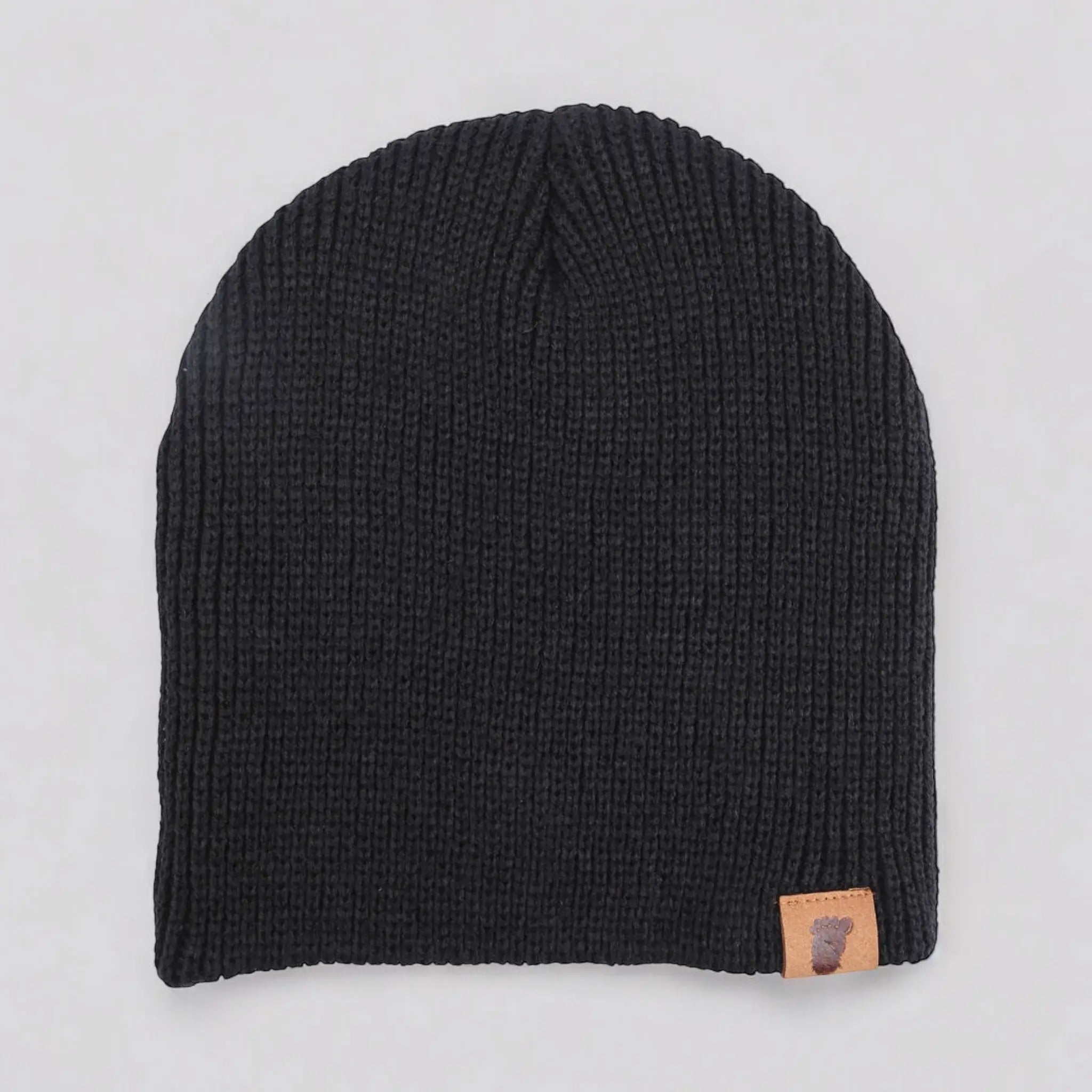Flat lay of the Wilderness Wanderer Beanie in Wood Booger Black — ribbed black knit beanie featuring leather Sasquatch footprint patch.