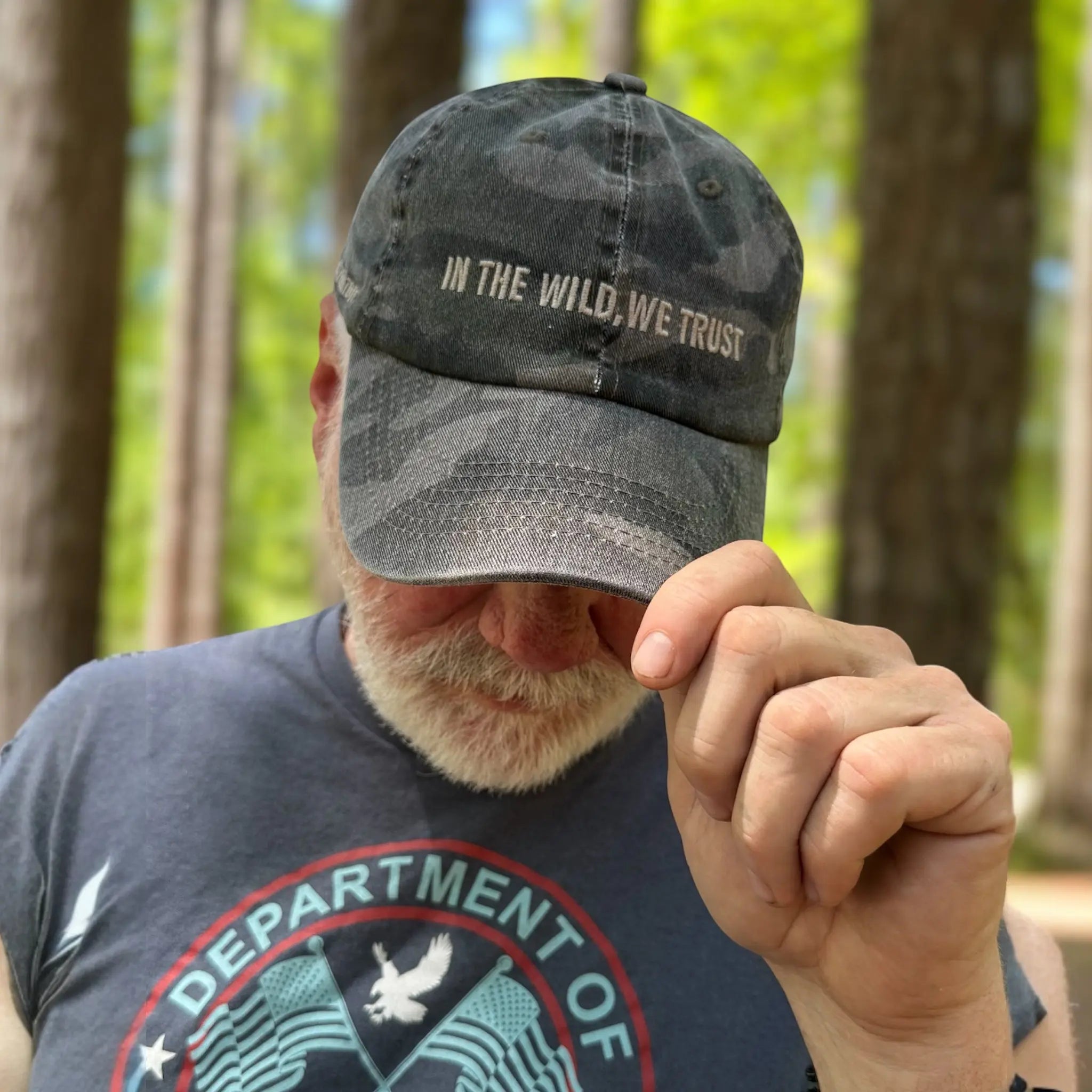 Man wearing Backwoods Camo Dad Hat in wooded setting; front text reads ‘In The Wild We Trust,’ perfect for outdoor wanderers and squatch chasers.