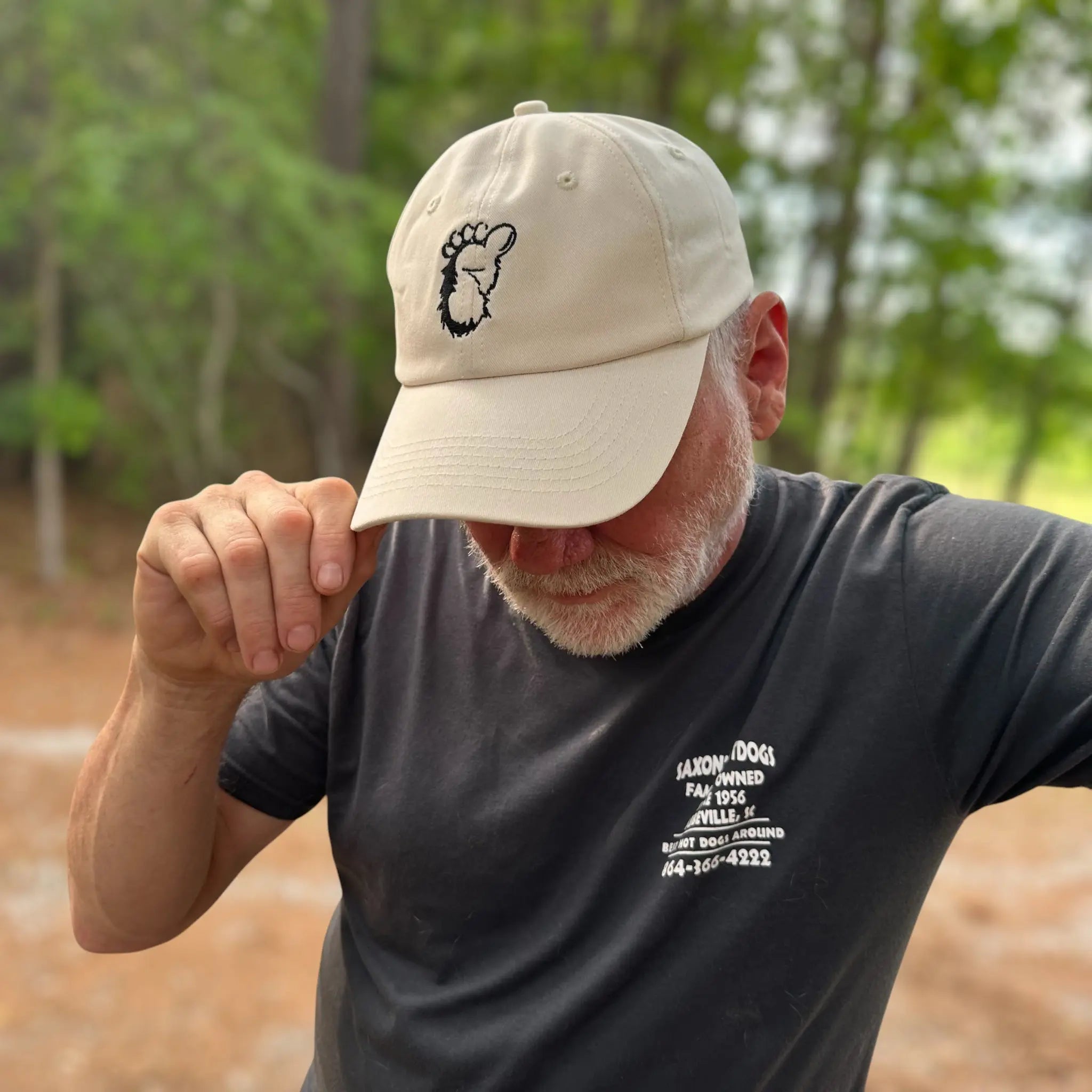 A man wearing the Sandstone Trail Dad Hat in the forest, gently tipping the brim. The neutral colorway and minimalist Sasquatch logo make it an ideal cap for camping, hiking, and wild-hearted adventurers.